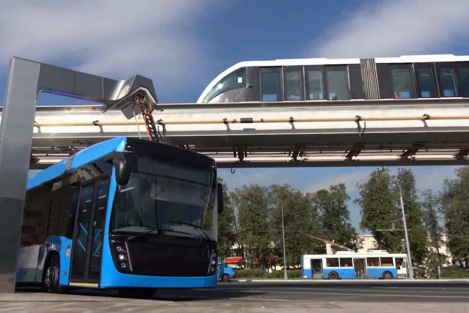Blue electric bus at charging station with monorail train passing on elevated rail above and older model trolley bus in background.